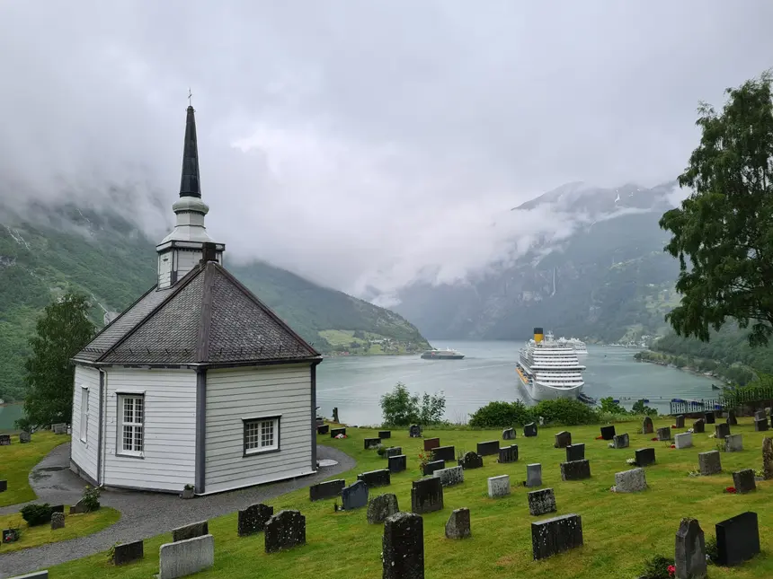 Achteckige weiße Kirche von Geiranger mit Friedhof, dahinter Kreuzfahrtschiffe im Fjord