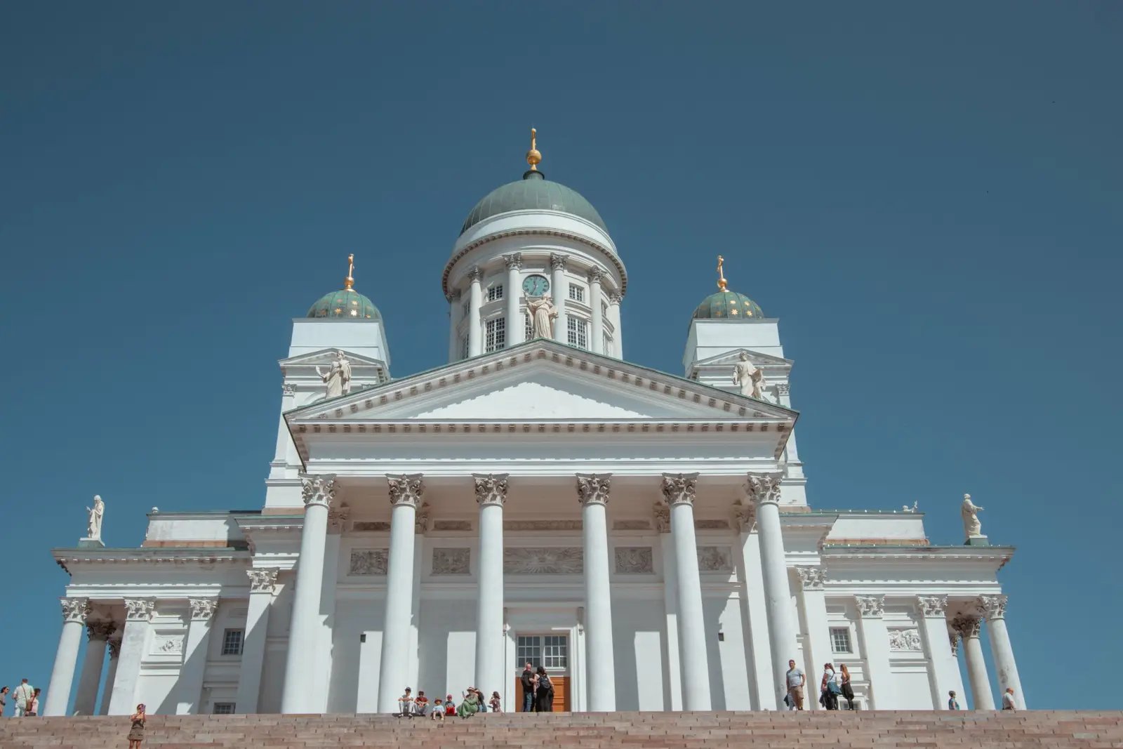 Strahlend weiße Säulenfront des Doms von Helsinki mit grünen Kuppeln vor blauem Himmel
