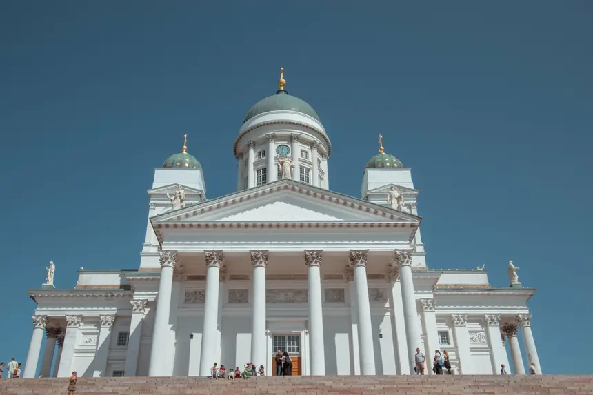 Strahlend weiße Säulenfront des Doms von Helsinki mit grünen Kuppeln vor blauem Himmel
