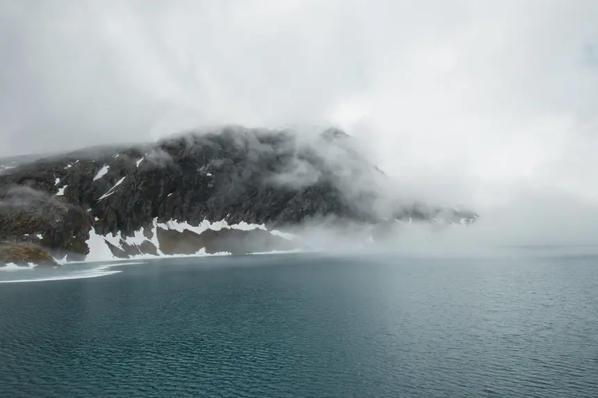 Nebel verschluckt einen schneebedeckten Berghang über dunklem Wasser