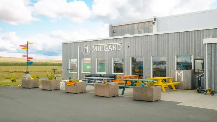 Das graue Holzgebäude des Midgard Base Camp mit Schriftzug, bunten Picknicktischen und Wegweiser davor